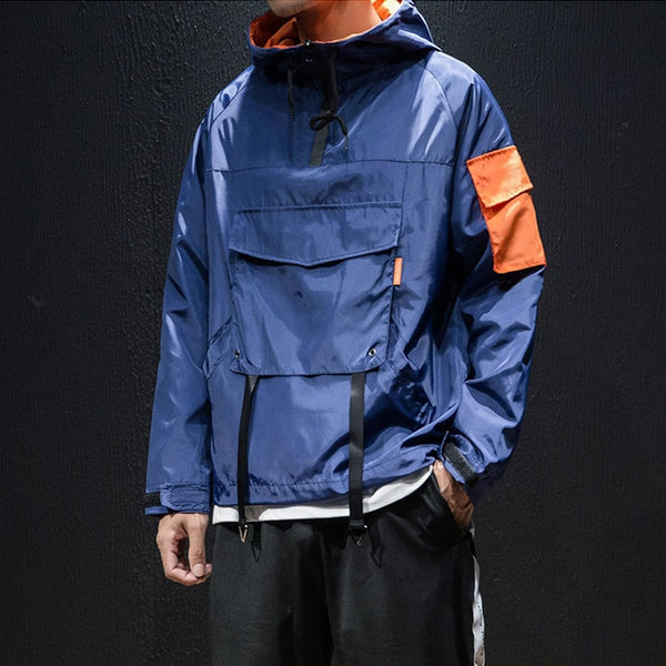 Un homme pose devant un fond noir. Il porte un jogging noir et une veste imperméable large bleu avec détails oranges et grande poche sur le devant.