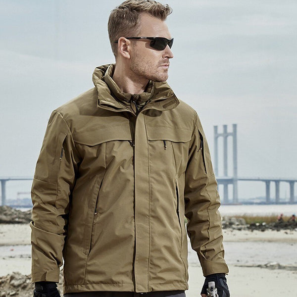 Un homme pose sur le sable devant un pont. Il porte un pantalon gris, des lunettes de soleil et une veste imperméable 3 en 1 avec doudoune légère amovible. La veste est beige. 