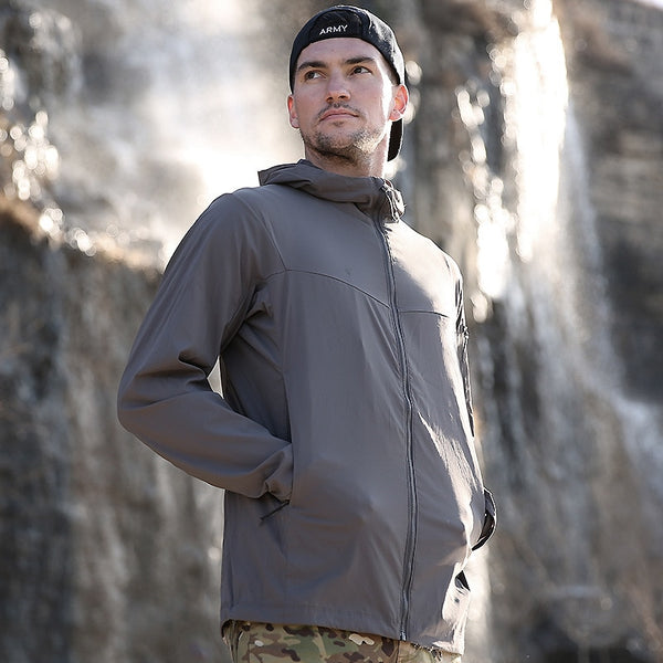 Un homme pose devant une cascade. Il porte une casquette noire, un pantalon vert camouflage ainsi qu'une veste imperméable anti UV respirant gris anthracite.