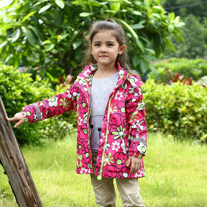 Une fille pose à l'extérieur. Elle porte un tee-shirt avec un ours, un pantalon beige et un manteau rose avec motif floral. 