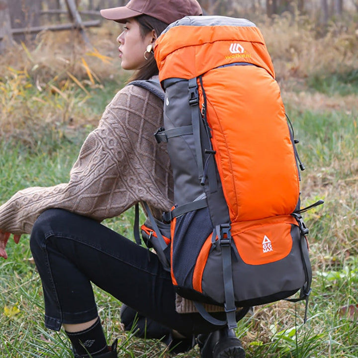 Une femme pose dans un champ accroupie. Elle porte un pull taupe en laine, un jean noir et des bottines. Elle porte une casquette marron et un sac à dos de camping orange et gris de 65L.