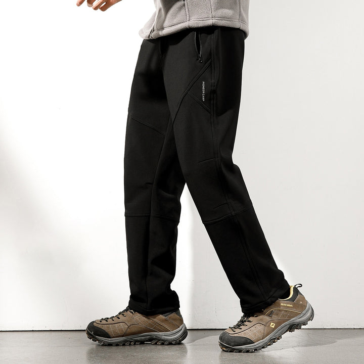 Un homme pose devant un mur blanc et un sol gris. Il porte une polaire beige, des chaussures basses de randonnée et un pantalon imperméable doublé polaire noir.
