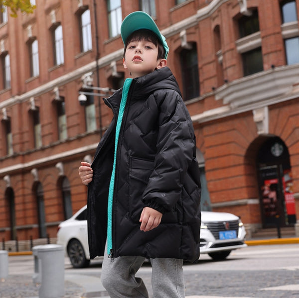 Un enfant pose dans la rue. Il porte une casquette bleu, un jean gris et une veste longue imperméable matelassée noir avec fermeture bleu.