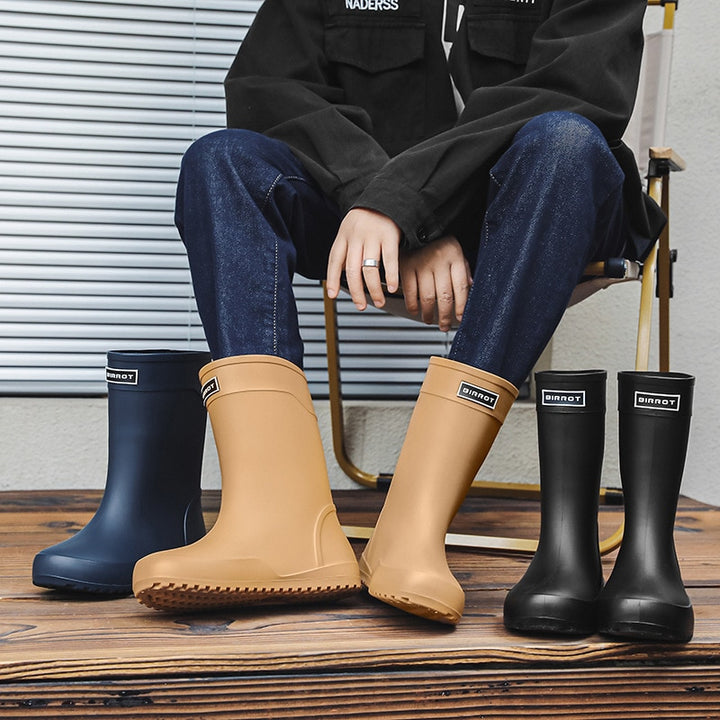 Un homme pose sur une terrasse en bois dans une chaise. Il porte une veste en jean noir, un jean brut ainsi qu'une paire de bottes de pluie imperméables beige. Il y a à côté une paire de bottes noires et une botte bleu marine.