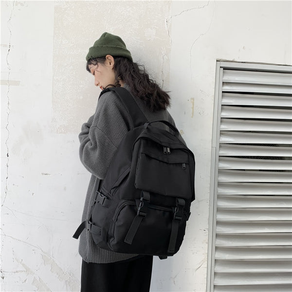 Une femme pose dans la rue devant un mur avec une grille. Elle porte un bonnet, un gilet, un pantalon noir ainsi qu'un sac à dos noir avec une grande poche sur le devant. 