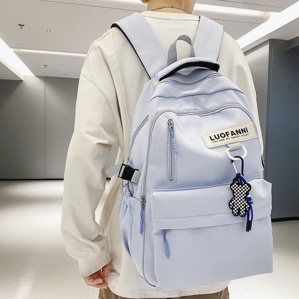 Un homme pose dans une salle vide aux murs gris. Il porte un tee-shirt long blanc, un pantalon beige et un sac à dos bleu ciel avec un pendentif damier accroché dessus.