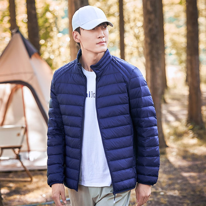 Un homme pose dans la nature, il y a une tente au fond. Il porte une casquette blanche, un tee-shirt blanc et un pantalon gris. Il porte une veste matelassée imperméable bleu. 