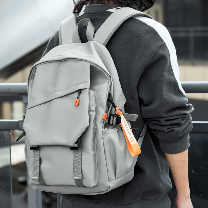 Un homme pose dans la rue avec un pull noir et gris. Il est de dos et porte un sac à dos gris clair qui a un rabat dans une former géométrique singulière.