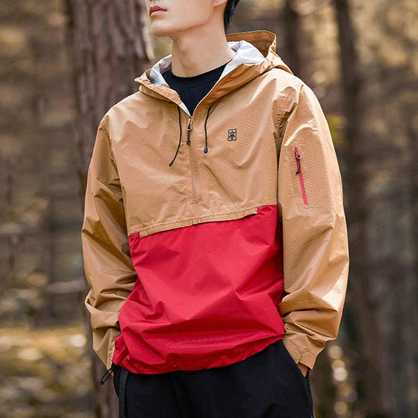 Un homme pose dans la forêt. Il porte un tee-shirt noir, un jean noir et une veste imperméable bicolore camel et rouge. 