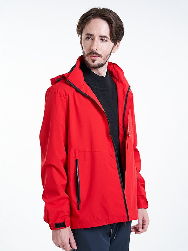 Un homme pose en veste imperméable coupe-vent coupe droite rouge. Il porte des vêtements noir et a des cheveux mi-longs bruns. 