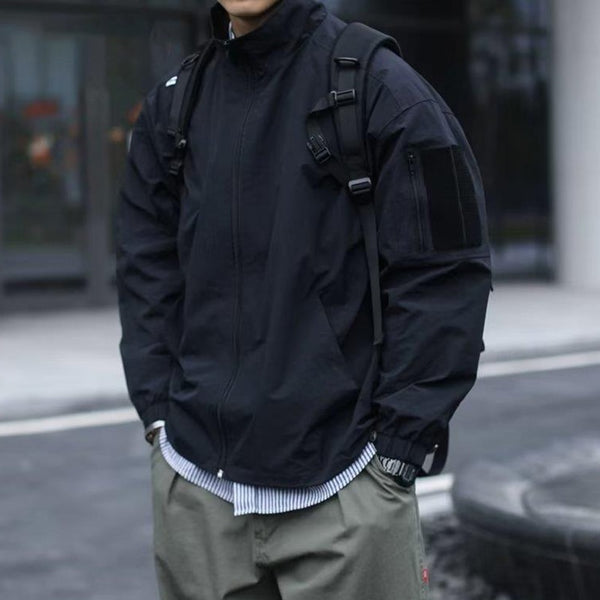 Un homme pose dans la rue. Il porte une veste imperméable coupe-vent noir large. Il porte aussi un sac à dos, une chemise rayée bleue et blanche ainsi qu'un pantalon gris.