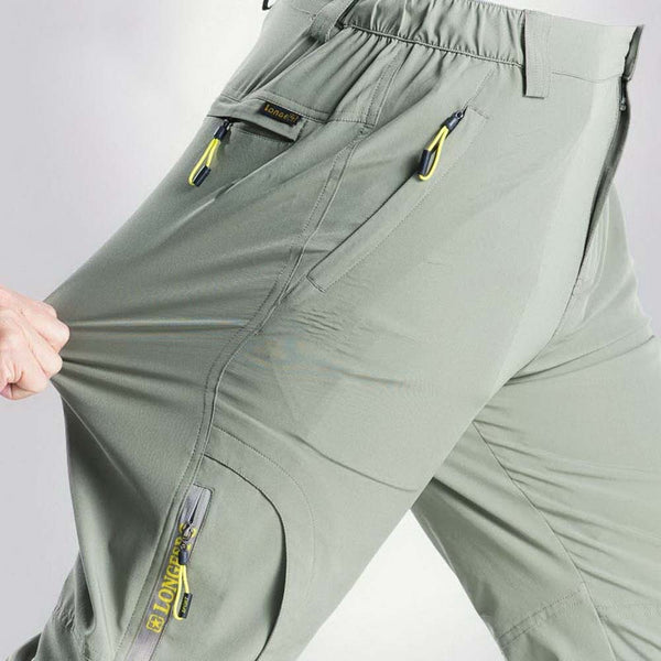 Un homme porte un pantalon de randonnée imperméable gris clair. Il a de nombreuses poches et un élastique à la taille. 