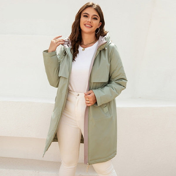 Une femme pose devant des murs extérieurs blancs. Elle porte un jean beige, un tee-shirt blanc et une veste mi-longue imperméable verte à capuche et doublure matelassée. Elle est brune aux yeux clairs.