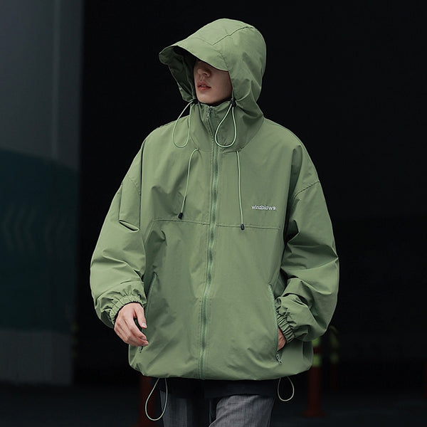 Un homme pose dans la rue. Il porte une veste imperméable vert large coupe-vent. Il porte aussi un jean.