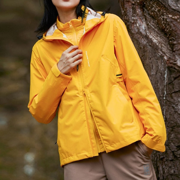 Une femme pose dans la forêt/ Elle porte une veste courte jaune imperméable et un pantalon marron.