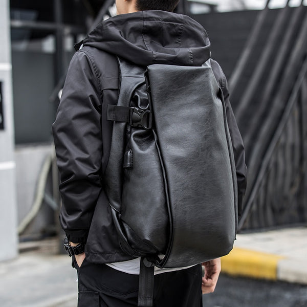 Un homme pose dans la rue. Il porte une veste à capuche noire, un pantalon noir, un tee-shirt blanc et un sac à dos en similicuir étanche imperméable avec port USB.