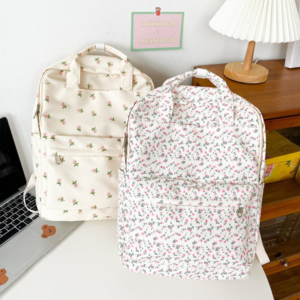 Deux sacs à dos blancs avec motif floral sont posés sur un bureau. Il y a un ordinateur à côté et une commode derrière. Sur la commode en bois il y a une petite lampe blanche.