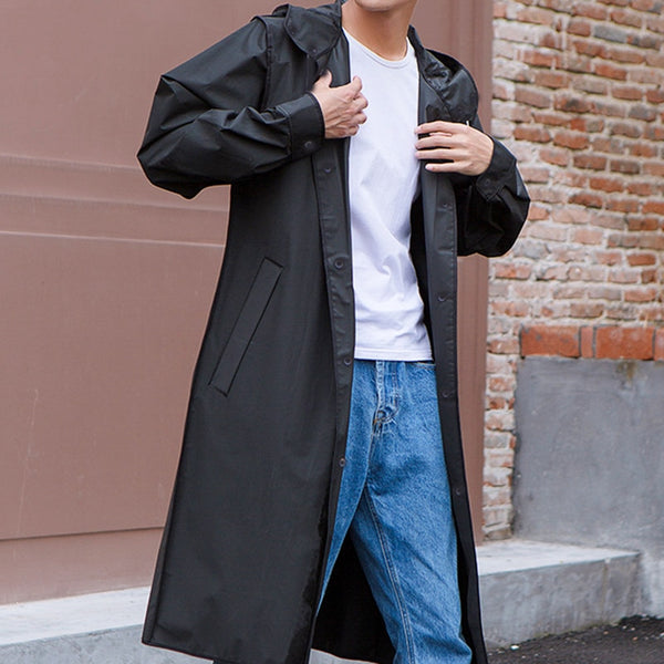 Un homme pose dans la rue devant un mur en brique. Il porte un tee-shirt blanc, un jean et une veste imperméable longue coupe-vent noir