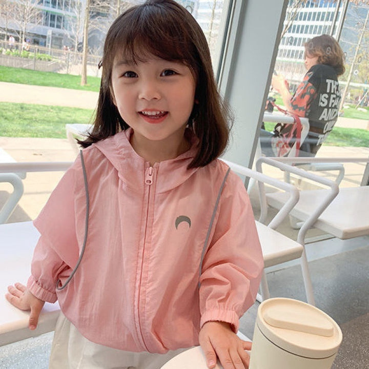 Une petite fille pose dans un café. Elle porte un pantalon blanc et une veste rose légère à capuche avec deux liserés roses et un croissant de lune gris. 