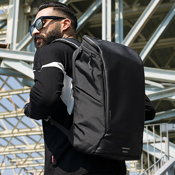 Un homme pose devant un pont en métal. Il porte des lunettes de soleil, un haut et un bas noir ainsi qu'un sac à dos imperméable et étanche au dos respirant. Le sac à dos est épuré et de couleur noire. 