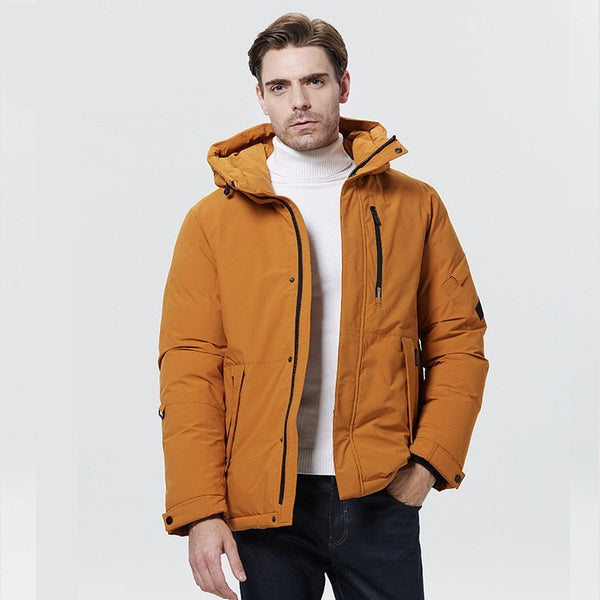 Un homme pose devant un fond gris. Il porte un col roulé blanc, un jean et une veste imperméable épaisse orange.