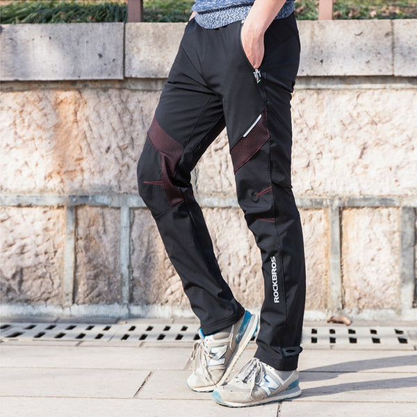 Un homme pose dans la rue avec des baskets grises et bleues, un tee-shirt bleu et un pantalon noir avec des patchs violets au niveau des genoux. Le pantalon est imperméable. 